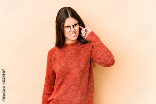 Young caucasian woman isolated on beige background covering ears with hands. © Asier