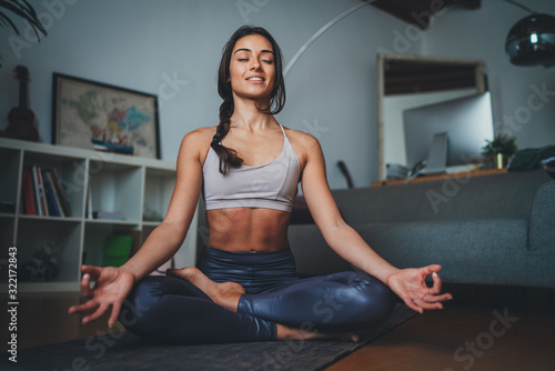 Young beautiful woman meditating at modern home interior sitting on yoga mat and smiling relaxed with closed eyes, Mindfulness meditation concept, Healthy Lifestyle