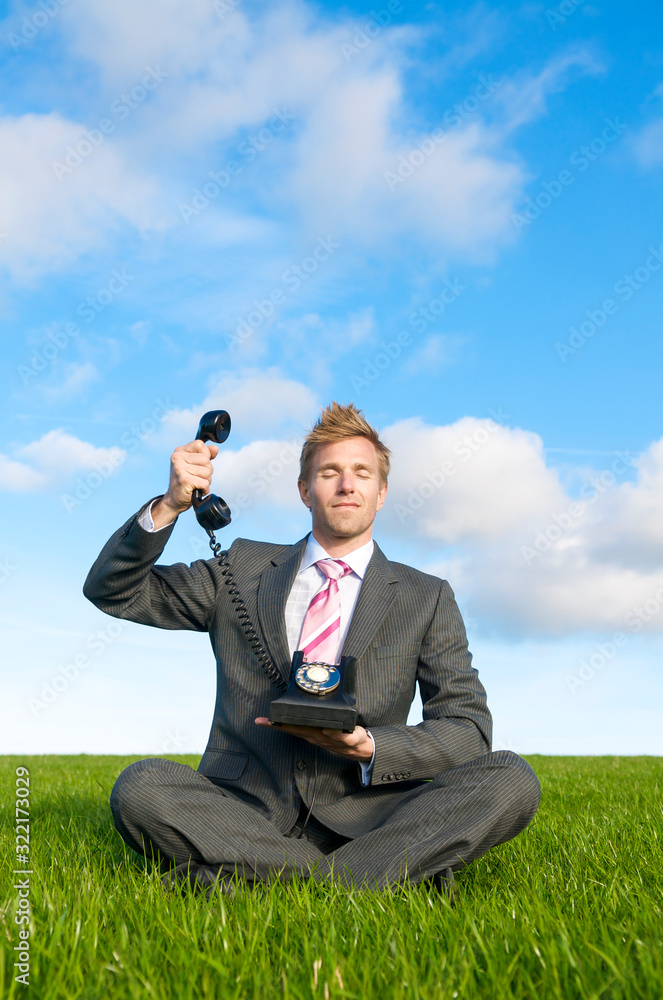 Calm businessman meditating in bright green meadow holding up the receiver of an old-fashioned rotary dial telephone Calm businessman meditating in bright green meadow holding up the receiver of an old-fashioned rotary dial telephone