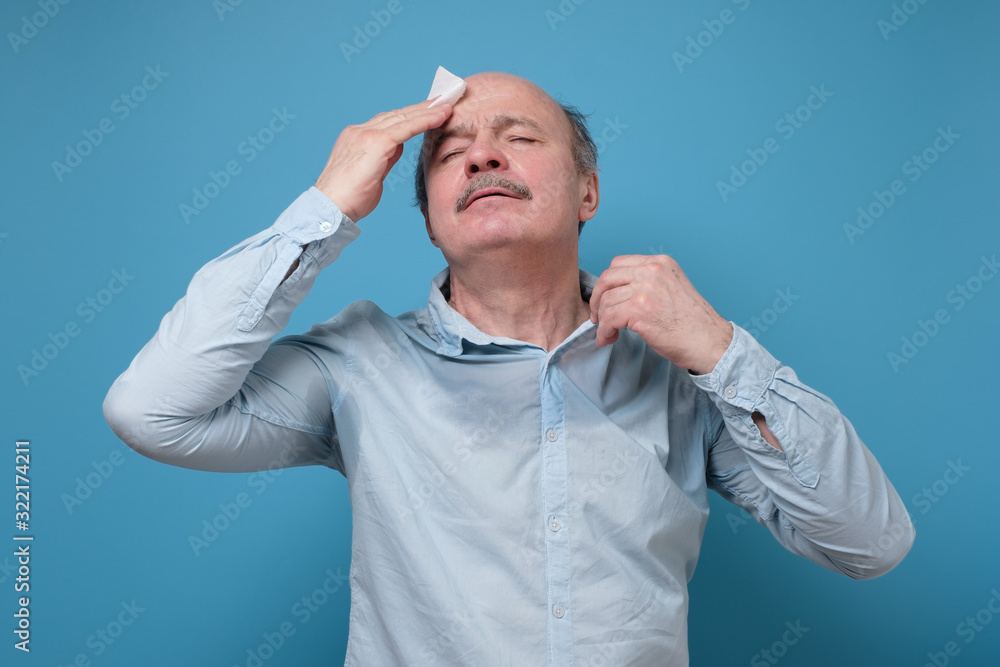Naklejka premium Senior hispanic man with sweat stain on his shirt suffering from hot weather. Studio shot on blue wall. Problem with hyperhidrosis