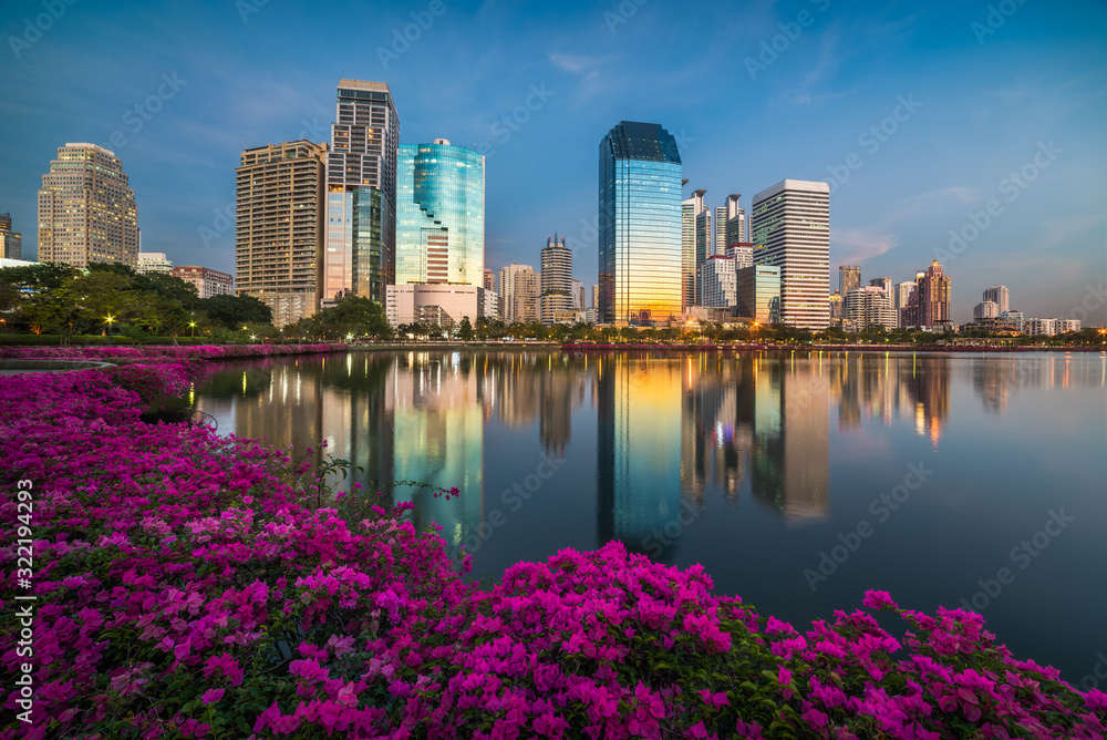 Fototapeta premium Lake with Purple Flowers in City Park under Skyscrapers at Twilight. Benjakiti Park in Bangkok, Thailand