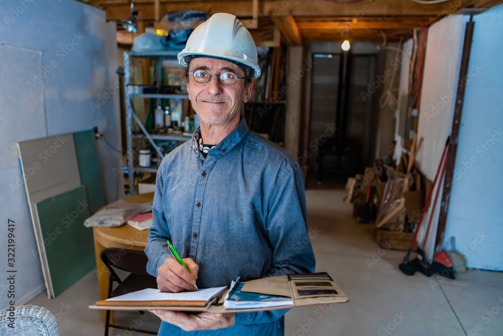 inspector wearing a safety helmet in the basement during a home inspection visit. A smiling middle aged caucasian man writes notes on notebook inspector wearing a safety helmet in the basement during a home inspection visit. A smiling middle aged caucasian man writes notes on notebook