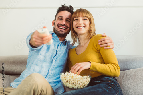 Happy beautiful couple watching TV and eating pop corn at home sitting on the sofa.