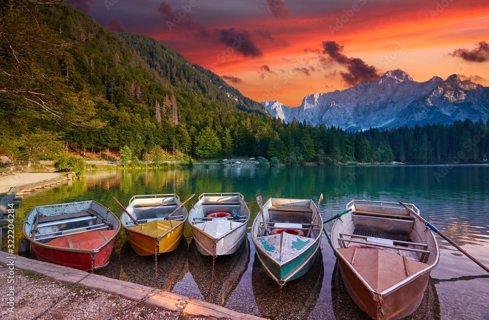 Amazing view on Lago di Fusine Inferiore at sunrise. Splendid morning scene of Julian Alps, Province of Udine, Italy, Europe. Beautiful red sky are reflected in the quiet lake. Five pleasure boats