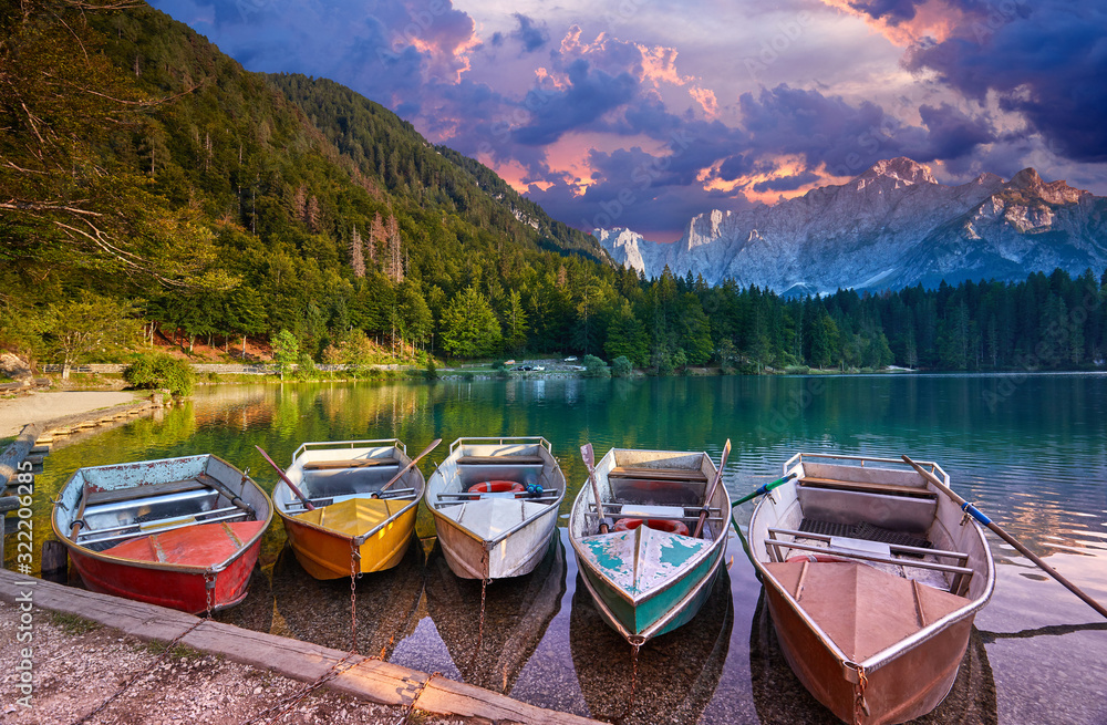 Amazing view on Lago di Fusine Inferiore at sunrise. Splendid morning scene of Julian Alps, Province of Udine, Italy, Europe. Beautiful forests are reflected in the quiet lake. Four pleasure boats