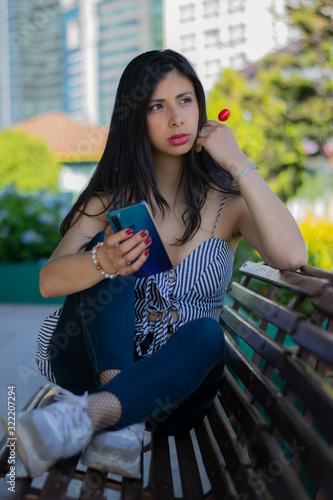 Colombian woman checks her cell phone sitting in a wooden chair eating a red candy