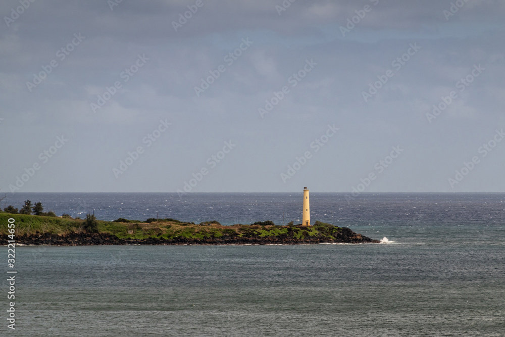 Nawiliwili, Kauai, Hawaii, USA. - January 17, 2020: Yellow Ninini lighthouse on black ocean-shore rocks of its green lands end. gray ocean water and light blue cloudscape.