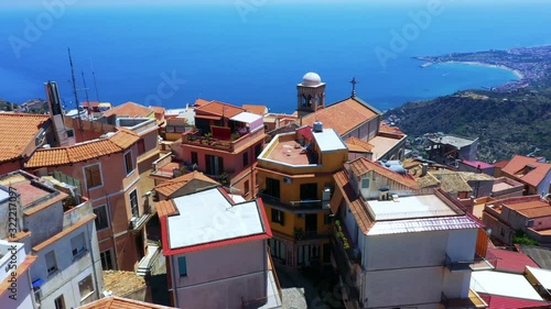 Aerial - drone flies over a small Sicilian village located on a cliff and reveals breathtaking view of green mountains with numerous houses and blue Mediterranean Sea in the background. Sicily, Italy. photo