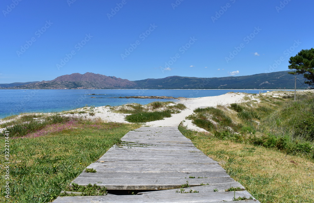 Beach with wooden boardwalk, sand dunes, mountain and bay with turquoise water and blue sky. Carnota, Spain.