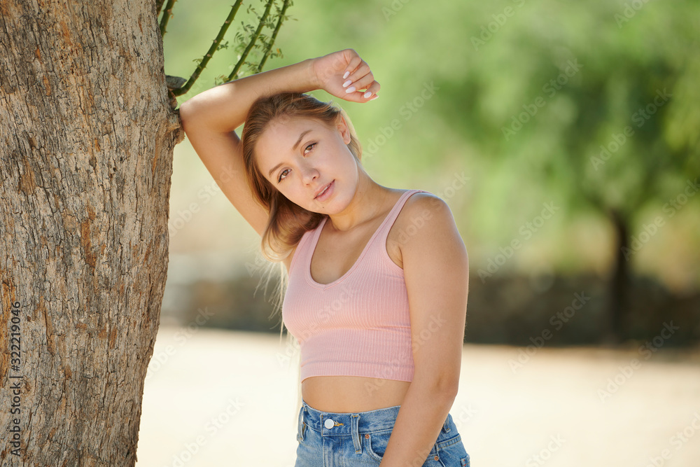 Attractive young Hispanic woman poses in desert under shade of tree wearing pink tank-top and denim shorts - leaning into tree