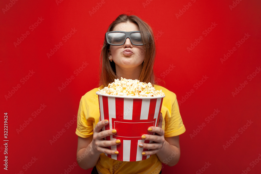 happy young girl the viewer in 3D glasses holds a big box of popcorn and watches a movie on a red color background happy young girl the viewer in 3D glasses holds a big box of popcorn and watches a movie on a red color background