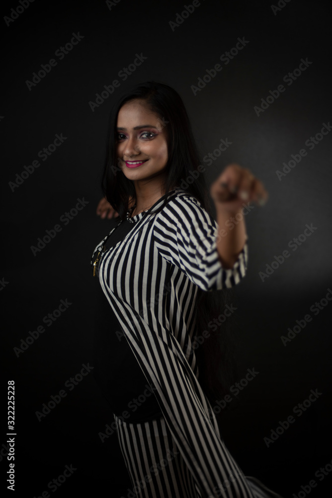 Obraz premium Fashion portrait of an young and attractive Indian Bengali brunette girl with striped western semi formal/casual dress in front of a black studio background. Indian fashion portrait and lifestyle.