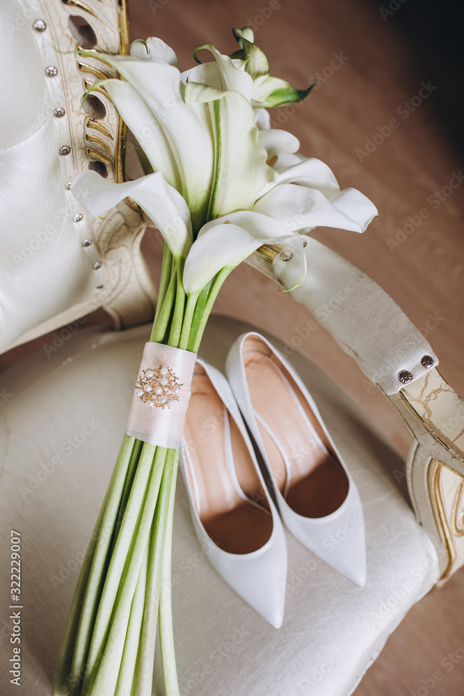 white flowers bride’s wedding bouquet with ribbon stands on a vintage chair next to high-heeled shoes
