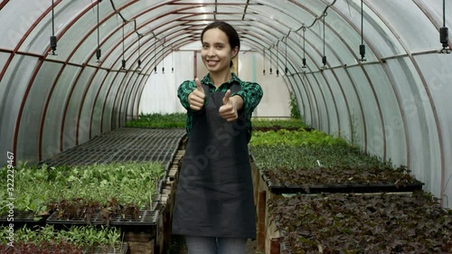 Young female farmer showing thumb up and smiling standing in the centre of greenhouse. Happy owner of agriculture small business is rejoicing at her success. photo