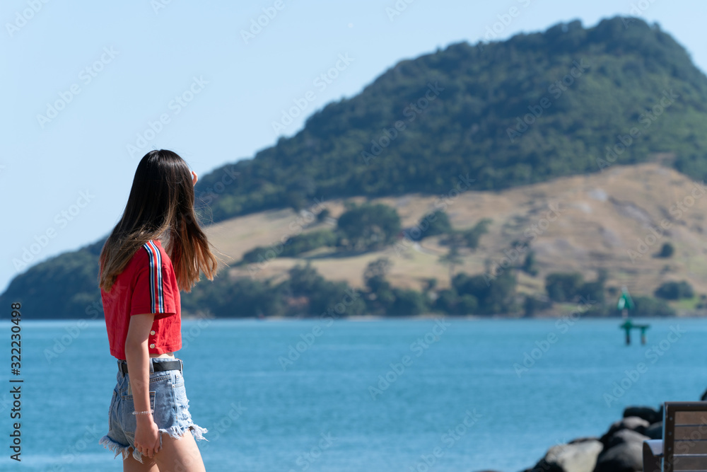 Teenage girl walking past  Tauranga Harbour with landmark Mount Maunganui in background on horizon.