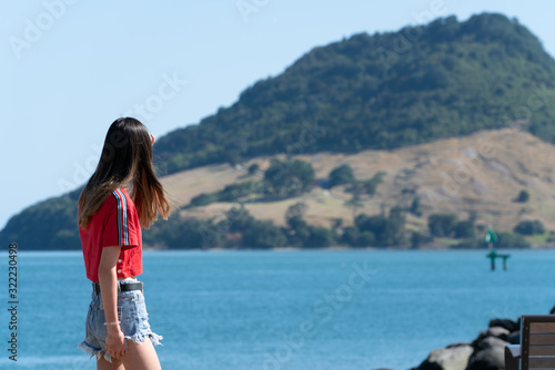 Teenage girl walking past Tauranga Harbour with landmark Mount Maunganui in background on horizon.