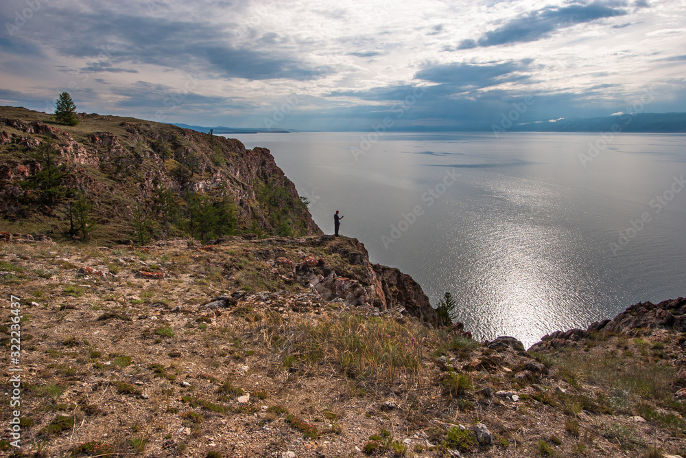 Large rock with a man on the edge. Lake Baikal. The sky in the clouds. Solar path on the water, ripples. On the rock trees.
