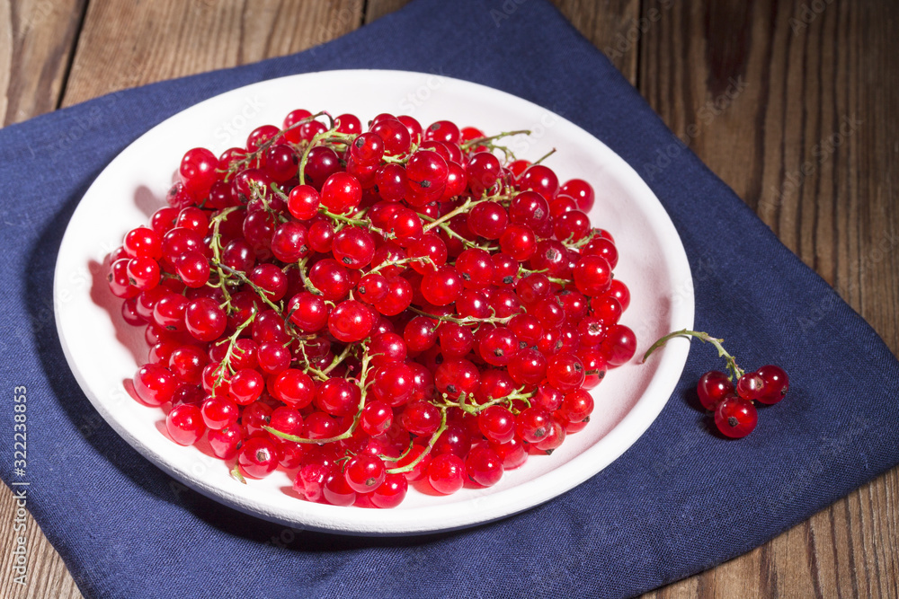 Fresh red currants in a white plate on a natural brown wooden background on line blue napkin. Close-up. Vegetarianism Fresh red currants in a white plate on a natural brown wooden background on line blue napkin. Close-up. Vegetarianism