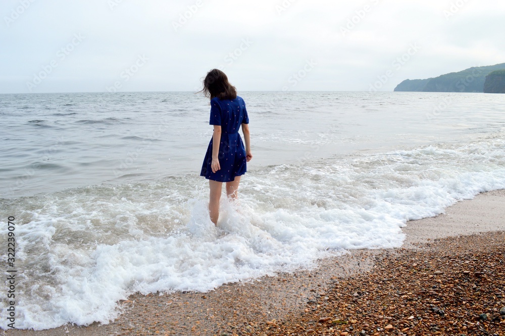 Upset thoughtful woman in blue dress going to the sea in cloudy weather before rain. She looking into the water