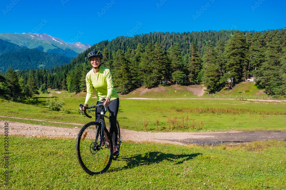 A young smiling girl on a cyclocross bike rides a green bright meadow against the background of a coniferous forest and a rural road