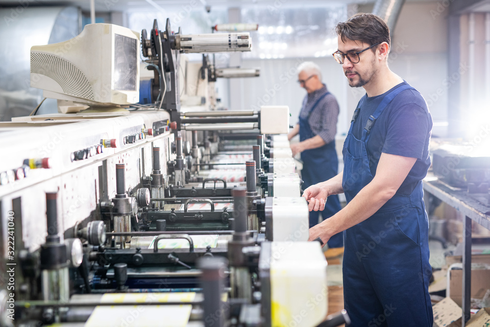 Serious young bearded employee of printing plant in blue overall working with modern machine
