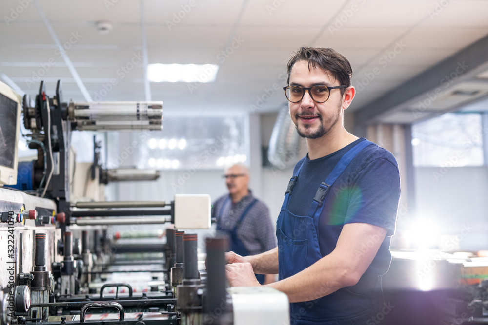 Portrait of content young printing specialist in glasses standing at printing press Portrait of content young printing specialist in glasses standing at printing press