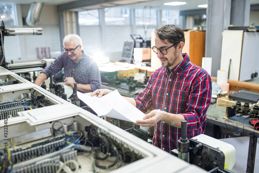 Smiling young worker in casual shirt standing at printing machine and checking printed paper Smiling young worker in casual shirt standing at printing machine and checking printed paper