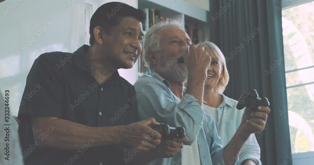 Group of senior people playing computer games in living room, competition. Caucasian, Asian, Thai people sitting on couch at home. Retirement lifestyle concept. Group of senior people playing computer games in living room, competition. Caucasian, Asian, Thai people sitting on couch at home. Retirement lifestyle concept.