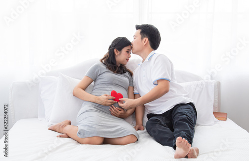Beautiful Asian pregnancy woman, husband hold belly, tummy with love, kiss, caring togetherness on bed at home, excited motherhood awaiting for childbirth sharing moment with couple, family concept