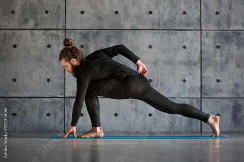 A young bearded man in black sportswear is practicing yoga against a gray wall.