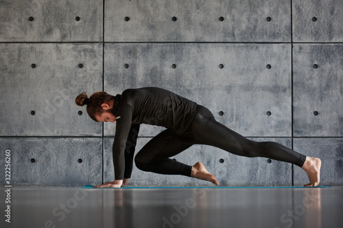 A young bearded man in black sportswear is practicing yoga against a gray wall.