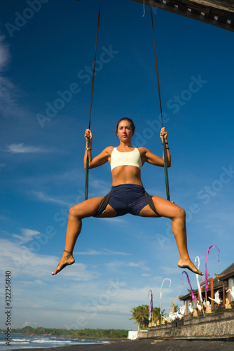 aerial yoga meditation beach workout - young attractive and healthy woman practicing aero-yoga training balance body and mind control hanging from ropes above the sea