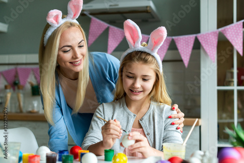 Happy Easter atmosphere! Funny, pretty mom making control, looking how her cute, small, joyful daughter draw, paint, decorate easter eggs, together wearing bunny ears