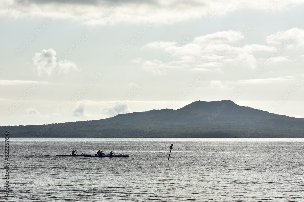 Crews paddling hard on board waka ama in calm channel in front of island with volcanic cone.