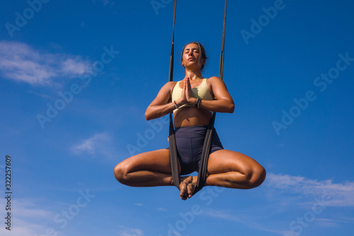 aerial yoga meditation workout  isolated on blue sky - young attractive and healthy woman practicing aero-yoga training balance body and mind control hanging from ropes