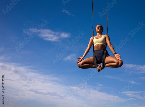 aerial yoga meditation workout isolated on blue sky - young attractive and healthy woman practicing aero-yoga training balance body and mind control hanging from ropes