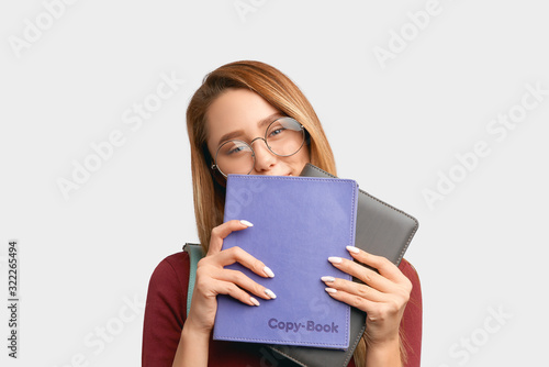 Student looks in camera hiding behind books. Beautiful young woman with long hair, clean smooth skin wears round large glasses for vision, backpack isolated on white background in Studio