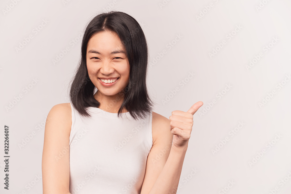 Girl showing gesture great thumbs up. Businesslike young woman Asian appearance with black hair and brown eyes dressed in short shirt stands isolated white background in Studio. Girl showing gesture great thumbs up. Businesslike young woman Asian appearance with black hair and brown eyes dressed in short shirt stands isolated white background in Studio.