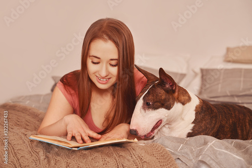 Fototapet Sweet tender young woman is lying in bed with bull Terrier in comfort of home apartment evening