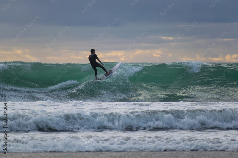 View of man surfer in black wet suit riding up the wave in evening light
