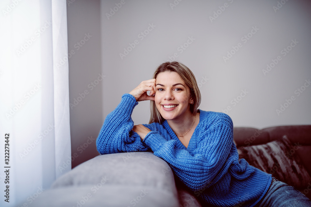 Cheerful caucasian blond young woman in sweater posing on sofa in living room and looking at camera. Cheerful caucasian blond young woman in sweater posing on sofa in living room and looking at camera.