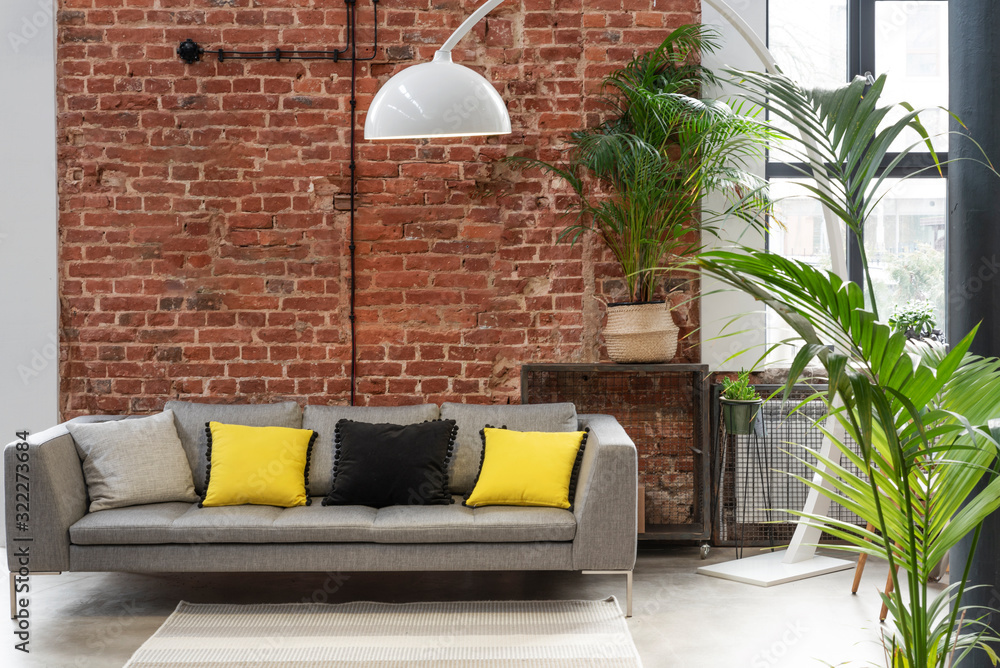 Living room interior in loft apartment in industrial style with brick wall,  grey stylish sofa with yellow pillows and big window. Modern lamp and  plants in minimal indoors. foto de Stock