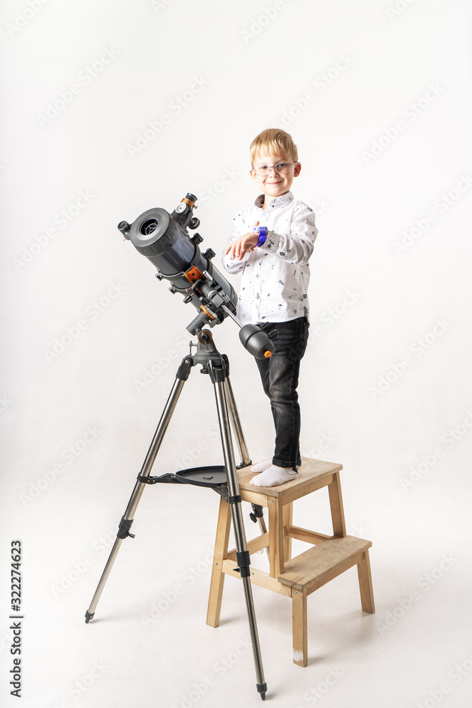 A little boy with glasses stands at a large reflex telescope in a chair.