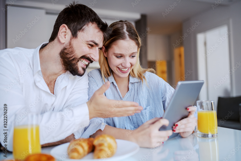Happy cheerful attractive caucasian couple sitting in dining room in the morning and looking at tablet. On dining table are glasses with juice and croissants.