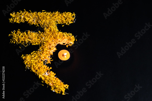 top view of tea lamp glowing with rupee sign made with flower petals on black background and copy space. financial concept. photo