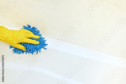 A female hand in a yellow rubber glove with a blue mop cloth washes and polishes the surface of a white bathtub. Spring  regular spring cleaning. Bathroom Cleaning Concept. Copy space for text.