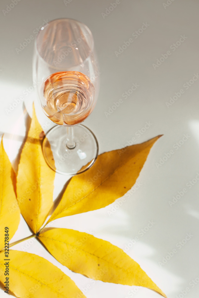 Glass of pink wine or fruit juice without pulp stands on a branch with bright yellow leaves. Simple minimalistic still life with an alcoholic drink on a white table. Sunny photo with shadows