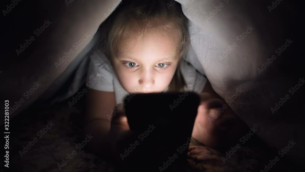 A young little girl is hiding under a blanket to use a digital tablet smartphone device late after sleep. Loneliness of young children and rescue on the phone. A young little girl is hiding under a blanket to use a digital tablet smartphone device late after sleep. Loneliness of young children and rescue on the phone.