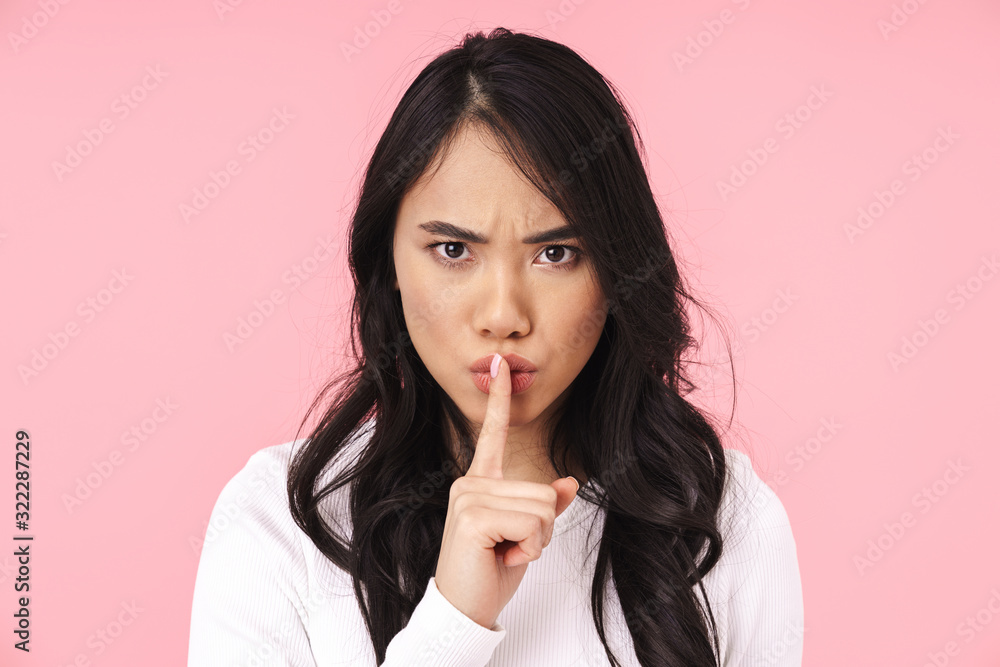Image of young brunette asian woman frowning and holding finger on lips Image of young brunette asian woman frowning and holding finger on lips