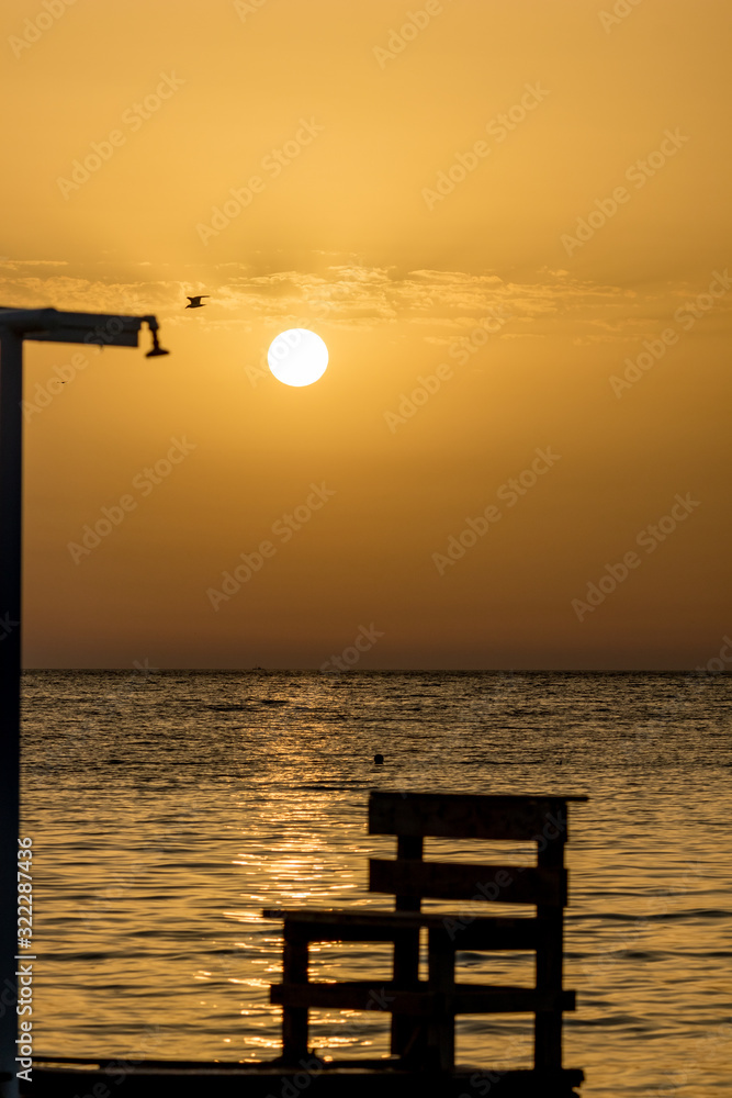 Focus on the round setting Sun. Blurred shower and bench silhouettes at the beach at sunset in front of golden Ionian Sea water near beach. Dusk as seen from Ksamil, Albania, spring scenery evening.